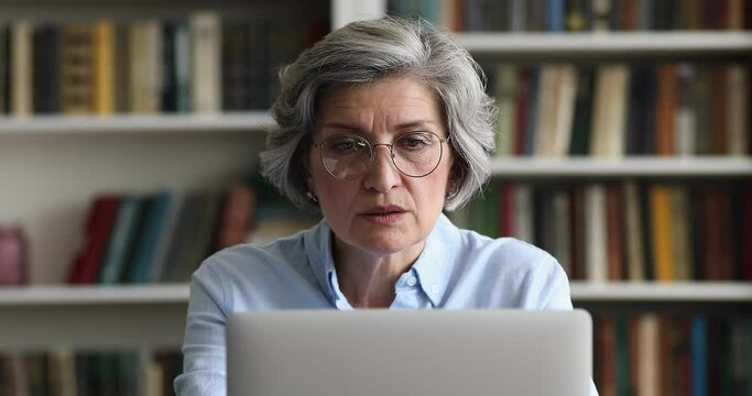 Close up smiling attractive older 50s businesslady in glasses working on laptop sit at desk in modern office look concentrated and motivated. Busy workday, workflow using modern tech, blogging concept