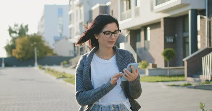 Close-up of attractive young caucasian woman walking on urban street and using modern smartphone outside, sunshine. Outdoors, lifestyle