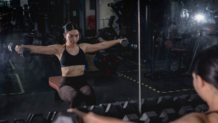 A determined and beautiful asian woman does a set of seated lateral dumbbell raises at the gym....