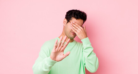 handsome hispanic man covering face with hand and putting other hand up front to stop camera,...