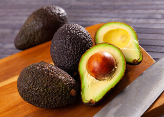 Whole and halved Hass avocados with typical dark green colored and bumpy skin on wooden cutting board with knife..