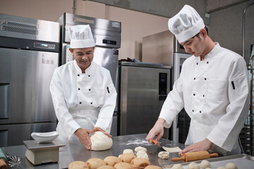 Two professional Asian male chefs in white cook uniforms and aprons are kneading pastry dough and eggs, preparing bread and fresh bakery food, baking in oven at stainless steel kitchen of restaurant.