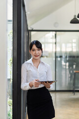 Obraz premium Asian woman holding a touchpad while standing in the office with large glass windows A female manager uses digital pads to organize her work.