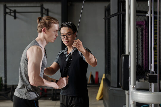 Fitness Trainer Correcting Muscular Young Man Doing Cable Pull Down Exercise For Tricep Muscles