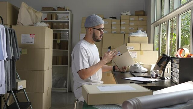 Asian male small business owner using mobile app on smartphone checking parcel box. Warehouse worker, seller holding phone scanning retail dropshipping package postal parcel bar code.