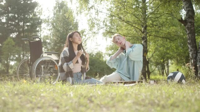 A disabled blonde man in a blue shirt and his girlfriend with dark long hair are making some healthy exercises in the public park, sitting on the plaid