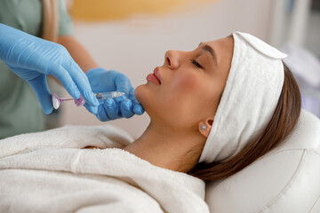 Side view of young woman getting hyaluronic acid injections in chin at beauty salon. Cosmetology - obrazy, fototapety, plakaty