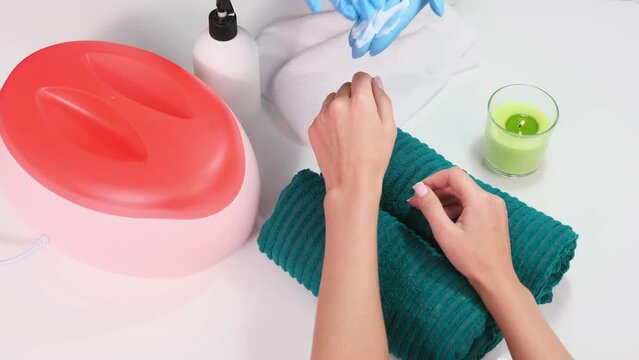 Master beautician in protective blue gloves taking some cream from the cream bottle and doing massage to a client hands with paraffin bath and candle on the table in beauty salon.