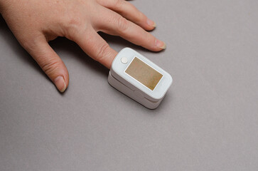 Fototapeta premium The oximeter is worn on the index finger of a woman's hand. A device for measuring blood oxygen levels and heart beats. Hands of an adult female against a gray background. Indecipherable readings.