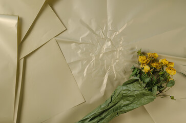 A bouquet of wilted flowers against a beige background. Dried yellow roses wrapped in green paper.