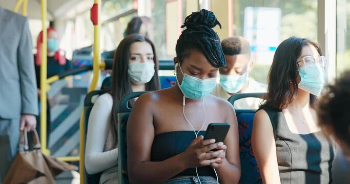 A beautiful woman with dreadlocks tied up in a bun and a protective mask on face is sitting on public transport bus smiling, looking at phone listening to music on her earphones