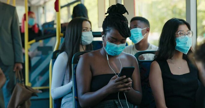 Pretty woman with dreadlocks tied up in a bun and a protective mask on face is sitting on public transport bus smiling, looking at phone listening to music on her earphones