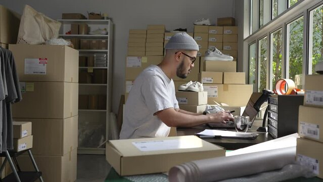 Asian man online seller confirming orders from customer on laptop. E-commerce business owner working on laptop computer in store warehouse.
