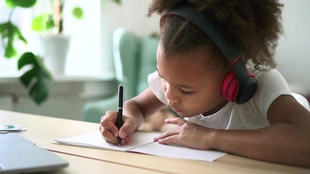 Little schoolgirl writing in notebook and sitting at table with laptop in home interior spbd. Closeup view of american african girl writes on paper and listens to lesson in headphones, studies