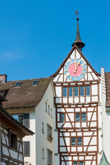Old dark brown medieval clock tower with cute weather vane on a white, half timbered tenement house with pastel colored and golden round clock on sunny day
