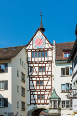 Fototapeta na wymiar Old dark brown medieval clock tower with cute weather vane on a white, half timbered tenement house with pastel colored and golden round clock on sunny day