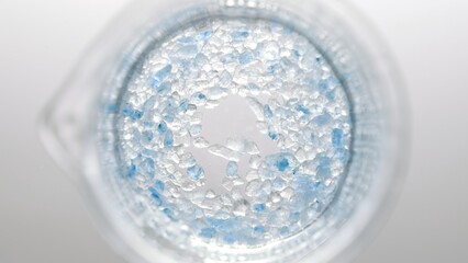 Top view of blue salt crystals dissolving in beaker with water on light grey background | Mineral skin care cosmetics formulation concept