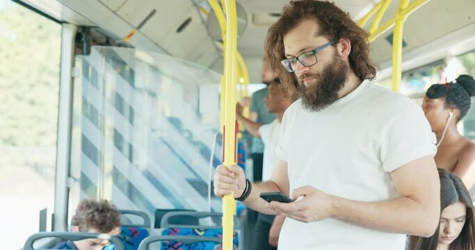 Young man with long hair and beard, wearing glasses, rides home in public transport vehicle, holds on to handrail, chats with girlfriend, sends messages on phone