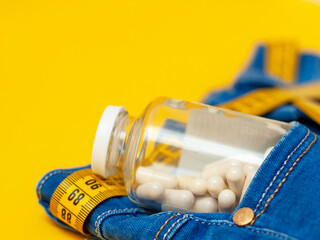 glass jar with pills in the pocket of blue jeans with a measuring tape on a yellow background, close-up, side view.