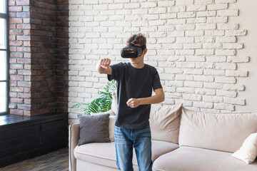 A teenage boy in a gray T-shirt and a virtual reality headset is boxing at home. Virtual sports and sports games in VR glasses. Hobbies and entertainment of teenagers