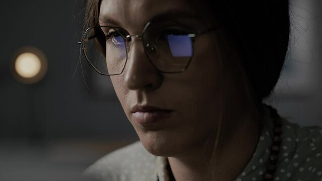 Freelancer typing message on laptop at night. Focused woman with glasses working at computer, screen is reflected in glasses. Close-up concentrated remote worker programmer fulfills order