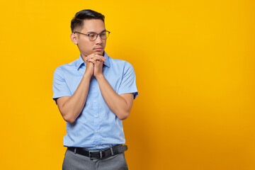 portrait of pensive young asian man in glasses keeps hand under a chin isolated on yellow background. businessman and entrepreneur concept