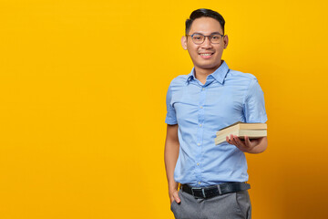 Fototapeta premium portrait of smiling young asian man in glasses holding book isolated on yellow background. businessman and entrepreneur concept