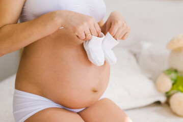 A woman with a big bare tummy is sitting on the bed holding baby booties in her hands. Pregnant lady strokes her belly. Tiny socks on a big belly