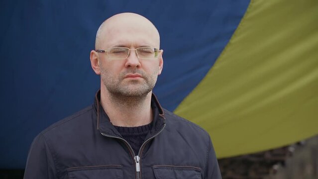 Serious refugee man standing near big waving Ukrainian flag in foreign country during invasion.Crisis, peace, stop aggression in whole world