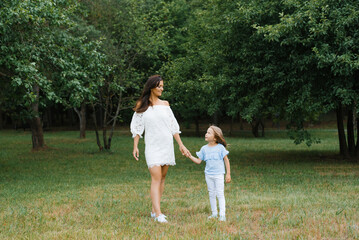 A young mother in a white dress holds her daughter's hand while walking in the park