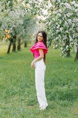 A cute young woman in white trousers and a bright pink blouse strolls through a spring park or garden among the flowering trees of an apple tree