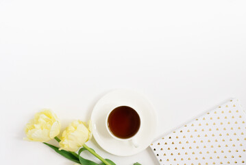 A white porcelain cup with black tea on a saucer surrounded by spring yellow tulips, a notebook on a white background with a copy space. The concept of spring, women's and mother's day, birthday 