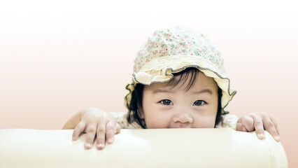 Close up of A little child is show her face standing behind the sofa. Portrait shot a little baby girl wear white hat is standing on the sofa. Little riding hood with smiling face is looking at camera