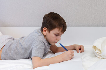 The child lying on the bed is engaged in lessons. The boy writes, draws with a pencil on paper. - 496759648