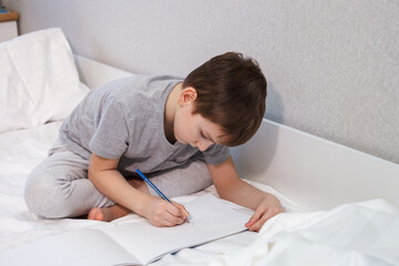 The child draws with a pencil on paper while sitting on the bed, close-up. Hobby