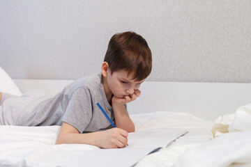 The child lying on the bed is engaged in lessons. The boy writes, draws with a pencil on paper. - 496759651