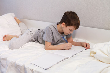The child lying on the bed is engaged in lessons. The boy writes, draws with a pencil on paper. - 496759652