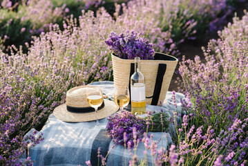 Two glasses with white wine and bottle on a background of a lavender field. Straw hat and basket with flowers lavender on a blanket on picnic table. Romantic evening in sunset rays. Time for dinner.