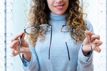 Close-up of a happy female client showing two glasses, making a choice. Stand of glasses in...