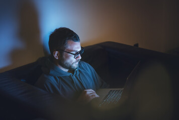 Young attractive serious man in glasses sitting at home at night on an armchair looking into a...