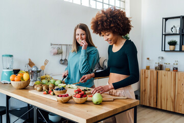 Smiling young woman cutting strawberries by friend eating apple at table in kitchen