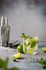 Mojito cocktail or mocktail with lime, mint, and ice in a glass on gray background. Summer cold alcoholic non-alcoholic drink, beverage and cocktail recipe. Bar menu. Copy space. Steel bar tools