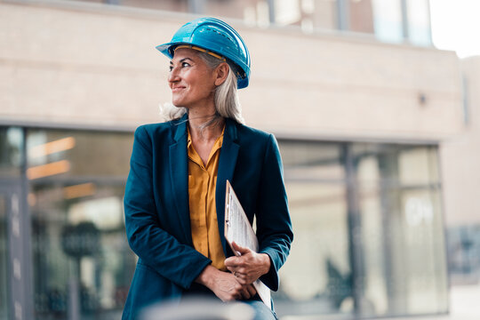 Smiling engineer wearing hardhat holding clipboard at office park