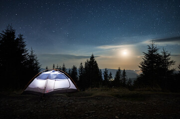 Fototapeta na wymiar Illuminated tent setting up on edge of stone trail on mountain hill. Incredible beauty of evening landscape. Moonlight, low clouds on background of starry sky. Mountain hills in the distance.