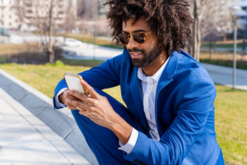 Businessman with curly hair using using phone sitting on footpath