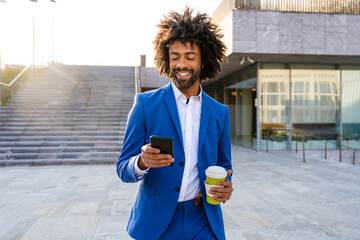 Smiling businessman holding disposable cup using mobile phone walking on footpath