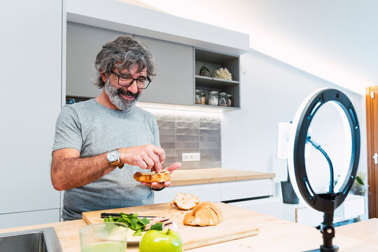 Smiling senior man making sandwich with croissant and vlogging through smart phone at home