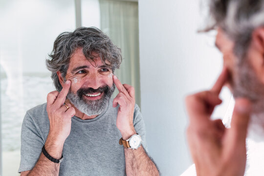 Smiling senior man looking in mirror and applying moisturizer on face
