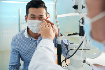 Asian man in medical mask complaining about pain in left eye when talking to ophthalmologist