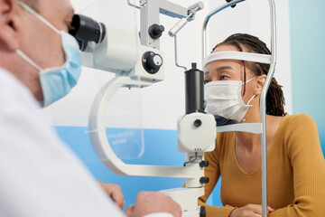 Black woman in medical mask getting eyes examined with autorefractometer to get lens prescription